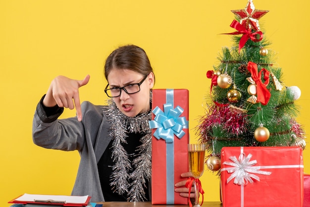 Vooraanzicht niet-gefeliciteerd meisje met bril zittend aan de tafel wees met vinger naar beneden kerstboom en geschenken cocktail