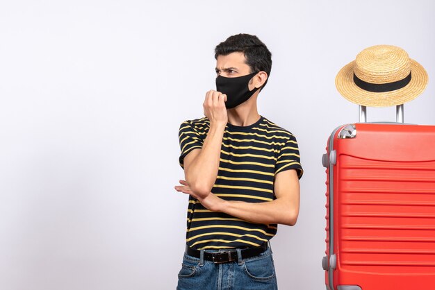 Vooraanzicht nieuwsgierige jonge toerist met zwart masker dat zich dichtbij rode koffer bevindt die iets ziet