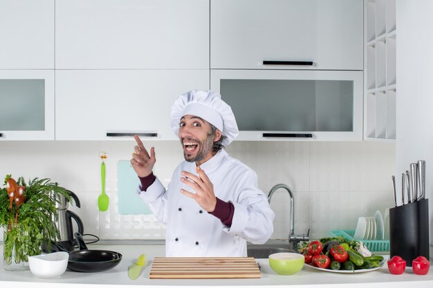 Vooraanzicht opgetogen mannelijke chef-kok die tong uitsteekt die achter de keukentafel staat