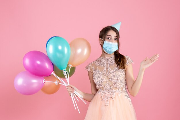 Vooraanzicht schattig feestmeisje met feestmuts die kleurrijke ballonnen houdt die naar iets wijzen