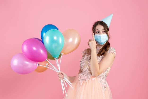 Vooraanzicht schattig feestmeisje met feestmuts die kleurrijke ballonnen houdt die vingerpistool maken