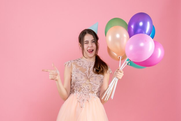 Vooraanzicht schattig feestmeisje met feestmuts met ballonnen wijzend met de vinger iets