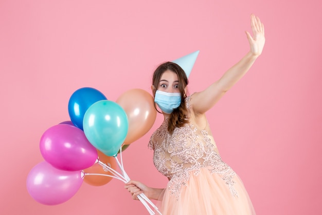 Vooraanzicht schattig feestmeisje met feestpet en medisch masker met kleurrijke ballonnen die haar hand opsteken