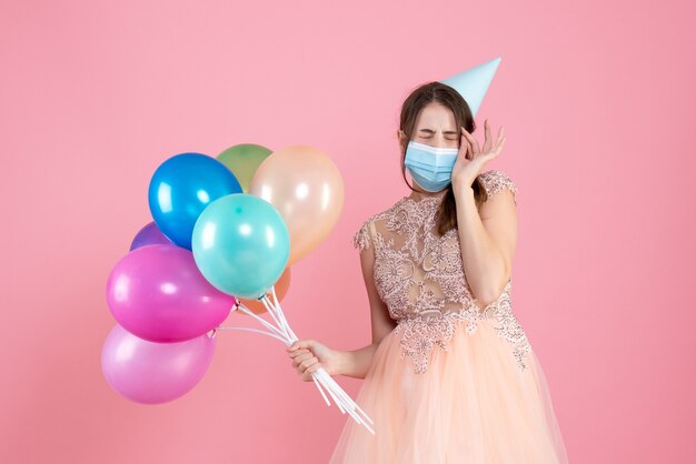 Vooraanzicht schattig feestmeisje met feestpet en medische masker sluitende ogen met kleurrijke ballonnen