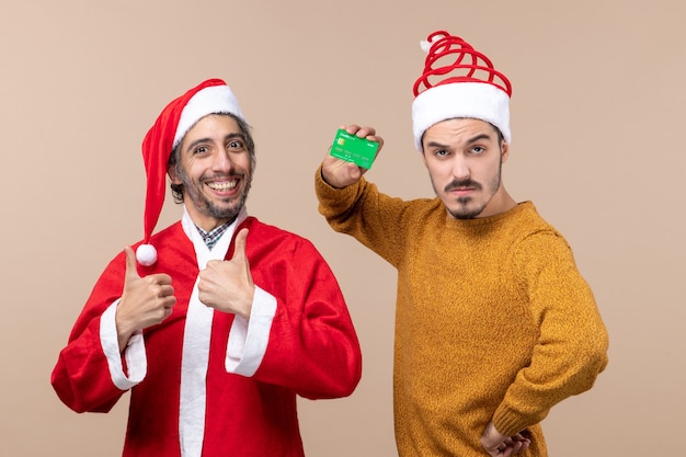 Gratis foto vooraanzicht twee gelukkige jongens één met santa vacht en de andere met krediet die zijn hand op een taille op beige geïsoleerde achtergrond zet
