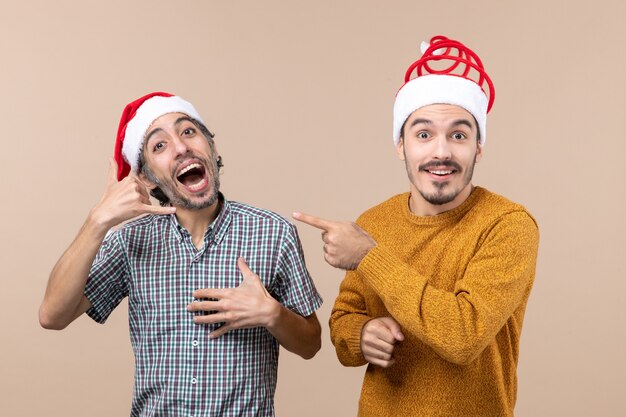 Vooraanzicht twee lachende jongens met kerstmutsen, een die bel me telefoonteken en de andere wijsvinger toont hem op beige geïsoleerde achtergrond