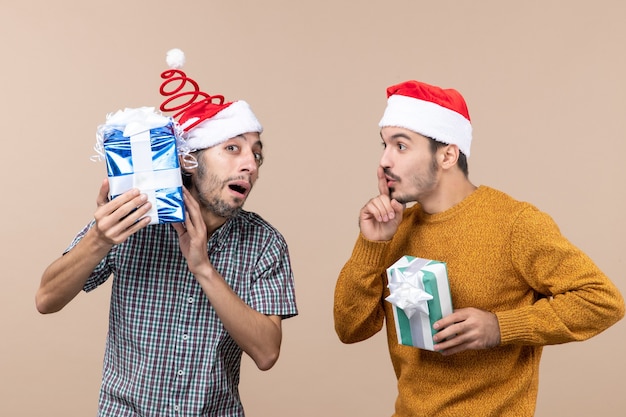 Vooraanzicht twee verwarde jongens die zijn kerstcadeautjes controleren en de andere shh-teken maken op beige geïsoleerde achtergrond