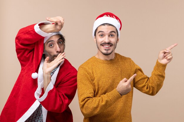 Vooraanzicht twee vrienden met santahoeden één die richting met gelukkig gezicht op beige geïsoleerde achtergrond tonen