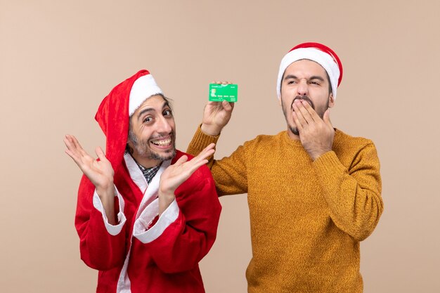 Vooraanzicht twee xmas jongens één met santa vacht en de andere met creditcard geeuwen op beige geïsoleerde achtergrond