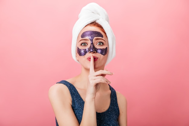 Vooraanzicht van aantrekkelijke vrouw die geheim teken toont terwijl het doen van spabehandeling. Studio shot van mooi meisje met gezichtsmasker geïsoleerd op roze achtergrond.