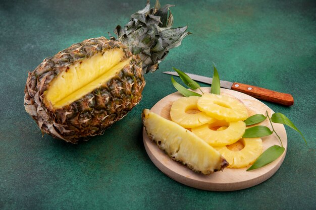 Vooraanzicht van ananas met een stuk uitgesneden van hele vruchten en ananas segmenten op snijplank met mes op groene ondergrond