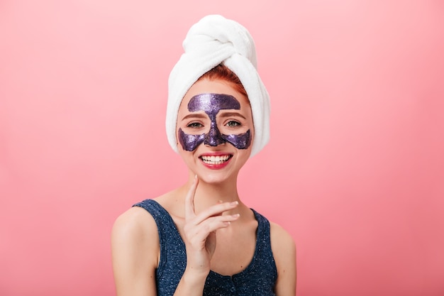 Vooraanzicht van betoverende vrouw met gezichtsmasker lachen op roze achtergrond. Studio die van zalig meisje met handdoek op hoofd is ontsproten die kuuroordbehandeling doet.