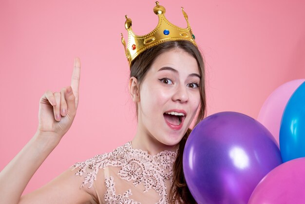 Vooraanzicht van de close-up schattig feest meisje met kroon houden ballonnen omhoog met vinger omhoog