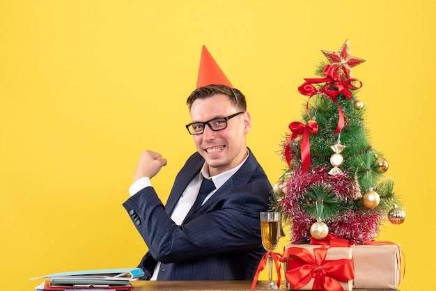 Vooraanzicht van de gelukkige zakenman met feestmuts zittend aan de tafel in de buurt van de kerstboom en presenteert op geel