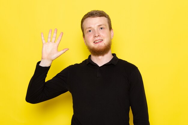 Vooraanzicht van de jonge man in zwart shirt met opgeheven hand en glimlach op gezicht