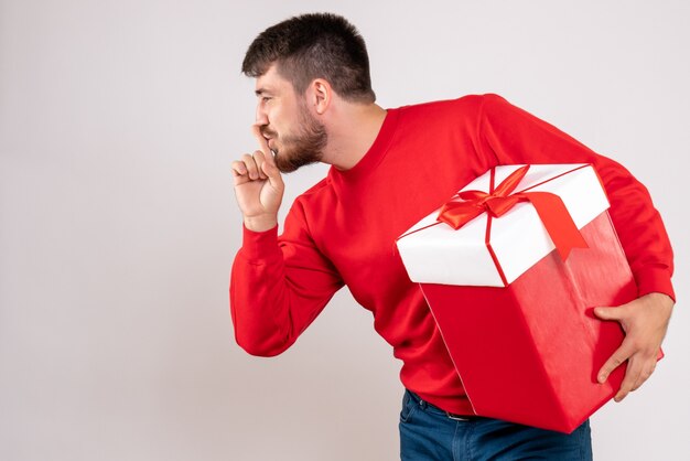 Vooraanzicht van de jonge mens die in rood overhemd kerstmis in doos op witte muur houdt