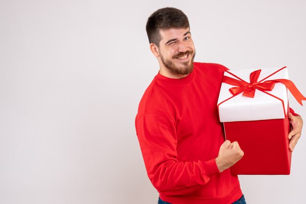 Vooraanzicht van de jonge mens die in rood overhemd kerstmis in doos op witte muur houdt