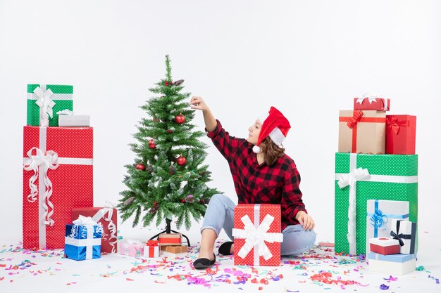 Vooraanzicht van de jonge vrouw die rond kerstcadeautjes zit en weinig vakantieboom op witte muur