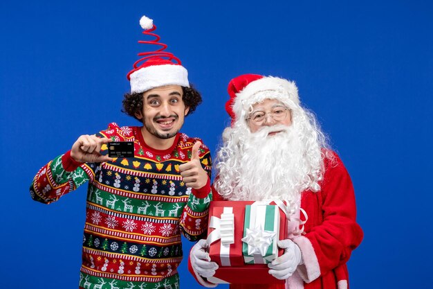 Vooraanzicht van de kerstman met jonge man met cadeautjes en bankkaart op de blauwe muur