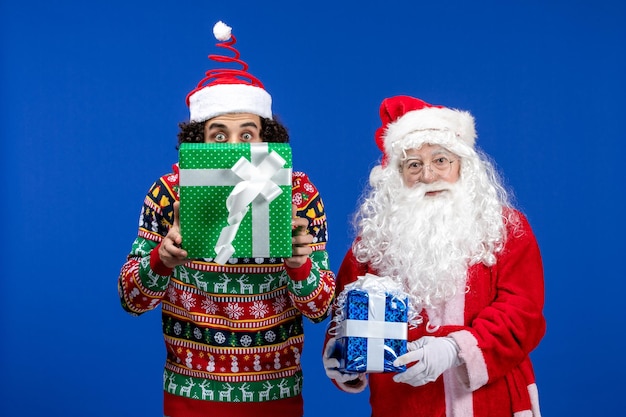 Gratis foto vooraanzicht van de kerstman met jonge man met kerstcadeautjes op de blauwe muur