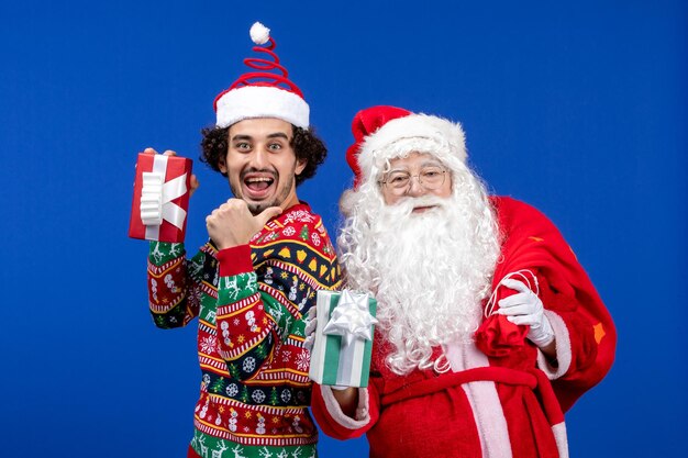 Vooraanzicht van de kerstman met jonge man met kerstcadeautjes op de blauwe muur