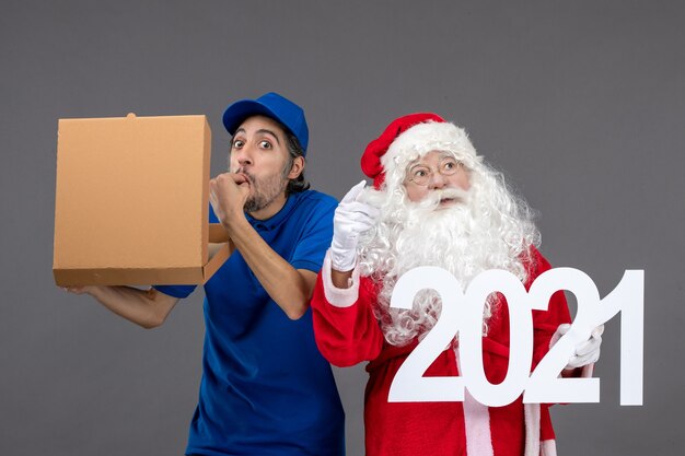 Vooraanzicht van de kerstman met mannelijke koerier met boodschappentassen en voedseldoos op de grijze muur