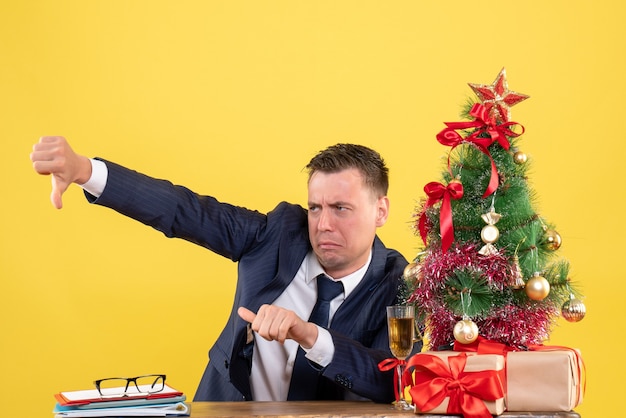 Vooraanzicht van de mens in pak die duim omlaag teken zittend aan de tafel in de buurt van kerstboom en geschenken op geel maken