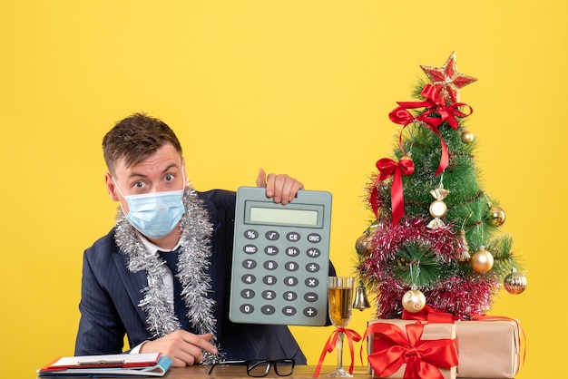 Vooraanzicht van de zakenman die calculator zittend aan de tafel in de buurt van de kerstboom en presenteert op geel