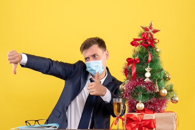 Vooraanzicht van de zakenman die duim op en neer teken zittend aan de tafel in de buurt van de kerstboom en presenteert op geel