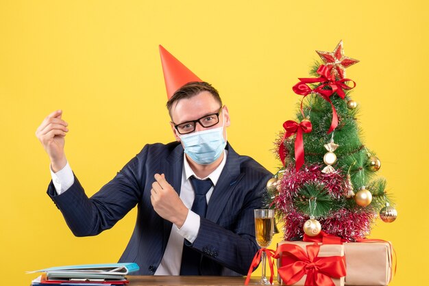 Vooraanzicht van de zakenman die geld maakt teken zittend aan de tafel in de buurt van de kerstboom en presenteert op geel