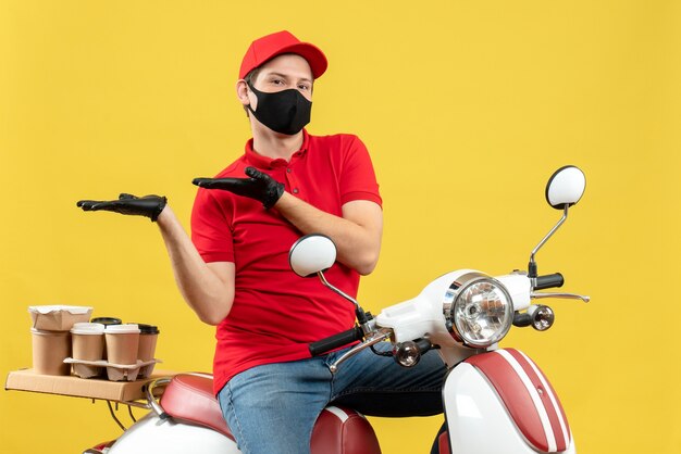 Vooraanzicht van denkende jonge volwassene die rode blouse en muts handschoenen draagt in medisch masker die bestelling levert zittend op scooter iets aan de rechterkant op gele achtergrond wijzend