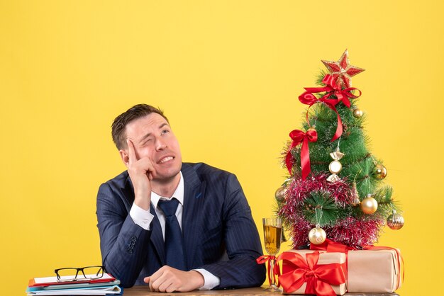 Vooraanzicht van denkende man met knipperende ogen zittend aan de tafel in de buurt van kerstboom en geschenken op geel