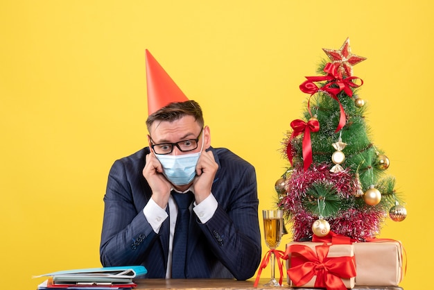 Vooraanzicht van depressieve zakenman zittend aan de tafel in de buurt van kerstboom en presenteert op geel