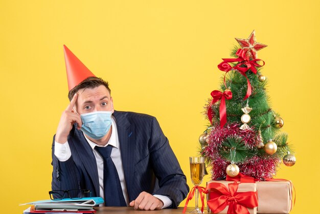 Vooraanzicht van doordachte man met feestmuts zittend aan de tafel in de buurt van kerstboom en presenteert op geel