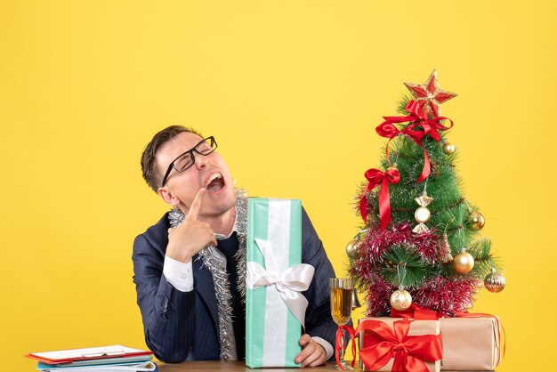 Vooraanzicht van doordachte man opening mond zittend aan de tafel in de buurt van kerstboom en presenteert op geel