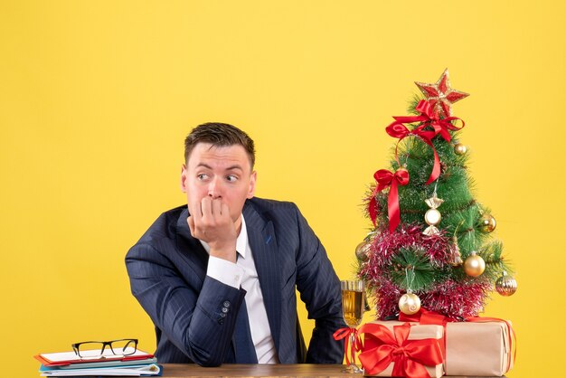 Vooraanzicht van doordachte man zit aan de tafel in de buurt van kerstboom en presenteert op geel
