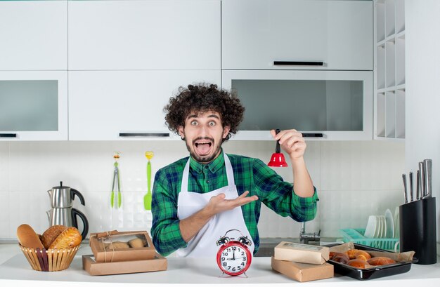 Vooraanzicht van een bange jongeman die achter de tafel staat met verschillende gebakjes erop en een rode bel in de witte keuken laat zien