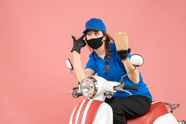 Vooraanzicht van een denkende bezorger met een medisch masker en handschoenen die op een scooter zitten en bestellingen afleveren op een pastelkleurige perzikachtergrond