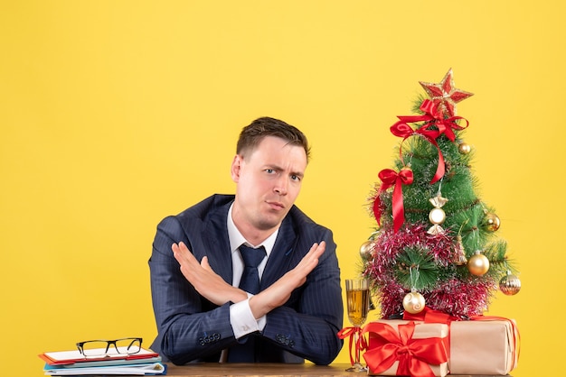 Vooraanzicht van een doordachte man die zijn handen kruist zittend aan de tafel in de buurt van de kerstboom en presenteert op geel