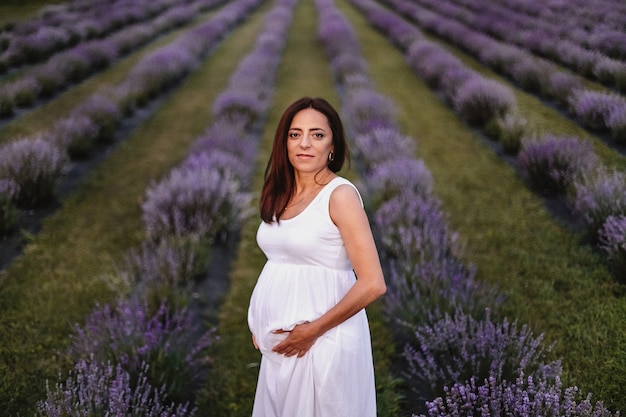 Vooraanzicht van een geglimlachte brunette blanke zwangere vrouw gekleed in witte jurk, buik aanraken in lavendelveld.