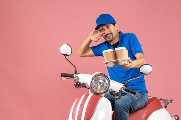 Vooraanzicht van een gekke, emotionele, grappige koeriersman met een hoed die op een scooter zit op een pastelkleurige perzikachtergrond