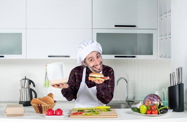 Vooraanzicht van een jonge hongerige man die een hamburger omhoog houdt die achter de keukentafel staat