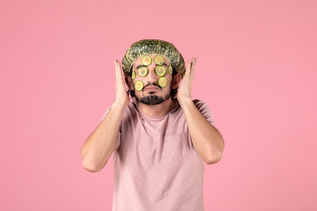 vooraanzicht van een jonge man die een komkommermasker op zijn gezicht op een roze muur aanbrengt