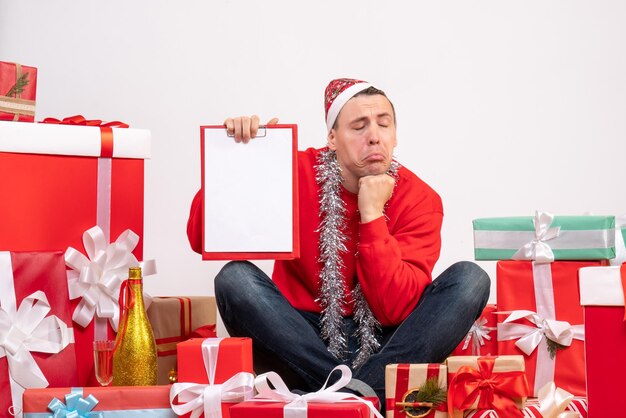 Vooraanzicht van een jonge man die rond kerstcadeautjes zit met klembord op een witte muur