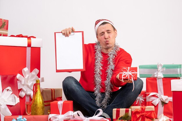 Vooraanzicht van een jonge man die rond kerstcadeautjes zit met klembord op een witte muur