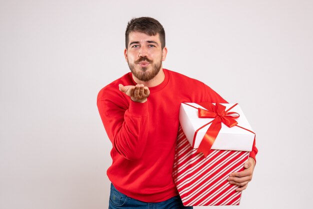 Vooraanzicht van een jonge man in een rood shirt met kerstcadeau in vak kussen verzenden op witte muur