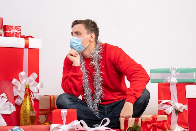 Vooraanzicht van een jonge man met een masker die rond kerstcadeautjes op een witte muur zit