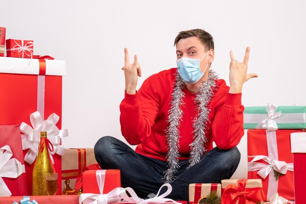 Vooraanzicht van een jonge man met een masker die rond kerstcadeautjes op een witte muur zit