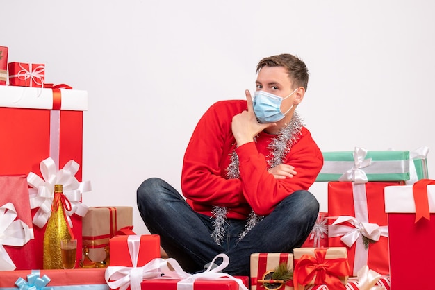 Vooraanzicht van een jonge man met een masker die rond kerstcadeautjes op een witte muur zit