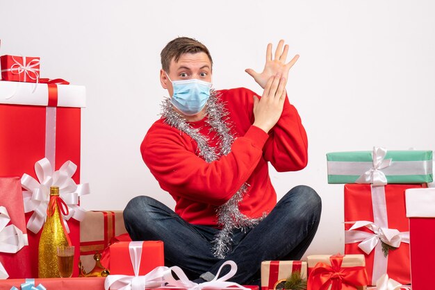 Vooraanzicht van een jonge man met een masker die rond kerstcadeautjes op een witte muur zit